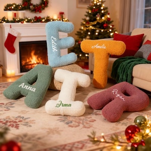 May include: A collection of plush, letter-shaped pillows in various colors, including green, blue, orange, white, and burgundy. Each pillow features a name embroidered in a contrasting color. The pillows are arranged on a patterned rug, with a Christmas tree and fireplace in the background.