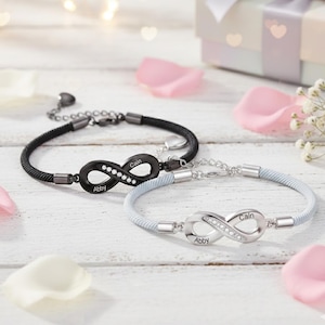 Puede incluir: Dos pulseras infinitas personalizadas. Una es negra con un dije infinito negro, la otra es plateada con un dije infinito plateado. Cada dije tiene los nombres "Cain" y "Abby" grabados y está adornado con pequeños cristales.