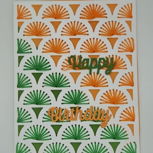Op de afbeelding: Handgemaakte verjaardagskaart met een witte achtergrond met een herhalend patroon van oranje en groene waaierachtige ontwerpen. De woorden "Happy Birthday" zijn uitgesneden en in oranje en groen op de kaart geplaatst.