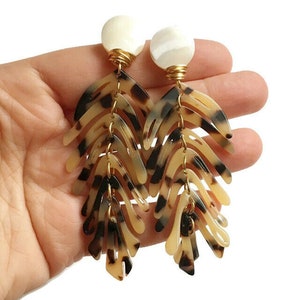 May include: A pair of long, dangling earrings with a tortoiseshell pattern. The earrings are made of multiple layers of plastic and have a white pearl at the top.