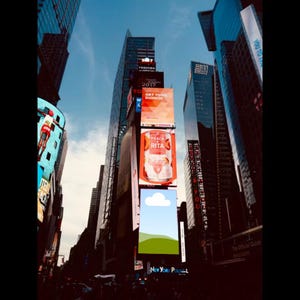 May include: A low-angle shot of Times Square in New York City, featuring towering skyscrapers and vibrant billboards. The billboards display advertisements for Dunkin' and Peach Rita, with the text "GET YOUR DUNKIN" and "PEACH RITA" visible. The sky is a clear blue.