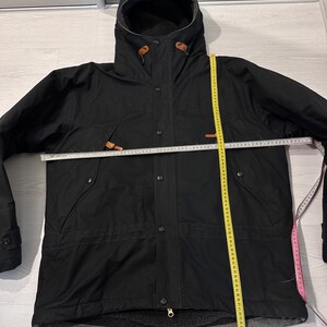 May include: A black hooded jacket with a front zipper and snap button closure, featuring brown leather-like toggles at the hood. A white measuring tape shows a width of approximately 26 inches, and a yellow tape shows a length of 30 inches.