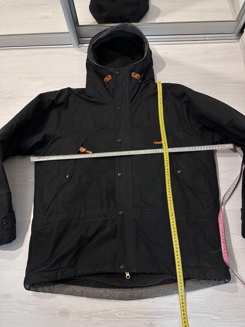 May include: A black hooded jacket with a front zipper and snap button closure, featuring brown leather-like toggles at the hood. A white measuring tape shows a width of approximately 26 inches, and a yellow tape shows a length of 30 inches.