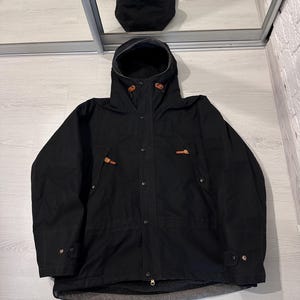 May include: A black hooded jacket featuring a front zipper closure with a snap-button placket, long sleeves with adjustable cuffs, and two chest pockets accented with brown leather toggles. The interior is lined with a dark gray fleece material.