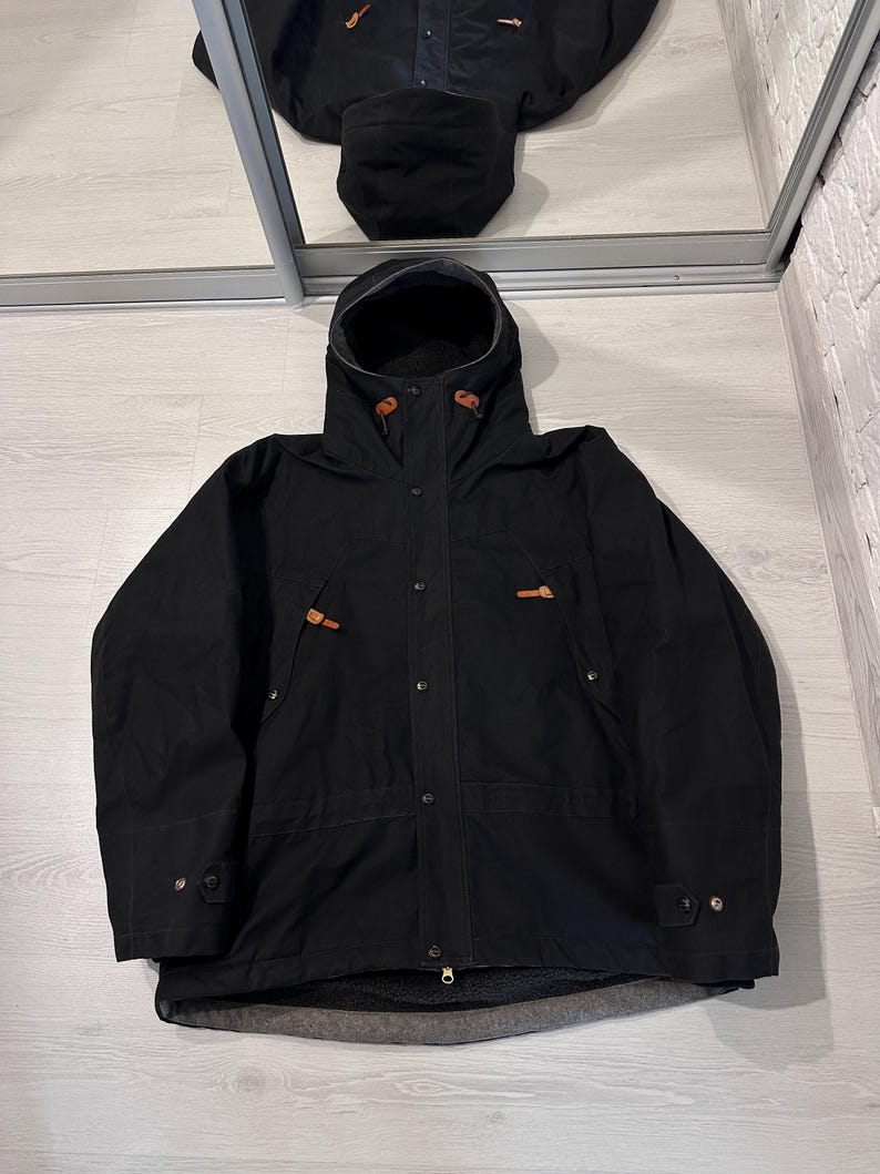 May include: A black hooded jacket featuring a front zipper closure with a snap-button placket, long sleeves with adjustable cuffs, and two chest pockets accented with brown leather toggles. The interior is lined with a dark gray fleece material.