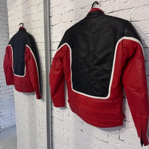May include: A red, black, and white leather jacket hangs on a black hanger. The jacket has a black upper back and shoulders, red sleeves and lower back, and white accents. The jacket is displayed in front of a white brick wall.