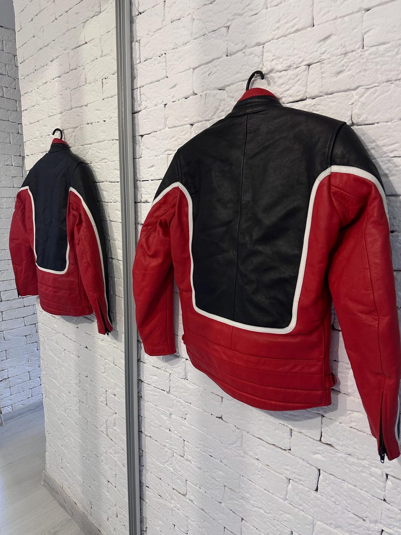 May include: A red, black, and white leather jacket hangs on a black hanger. The jacket has a black upper back and shoulders, red sleeves and lower back, and white accents. The jacket is displayed in front of a white brick wall.