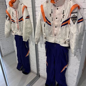 May include: A white ski jacket with orange and navy blue accents, featuring a high collar, front zipper, and WINDEXPRO branding on the sleeve, paired with matching solid navy blue ski pants that have orange and white geometric side details.
