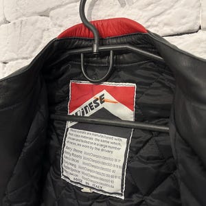 May include: Black leather jacket with a red collar, hanging on a black hanger. The jacket has a quilted black lining and a label with the brand name and text. The label also lists the names of World Champion riders.