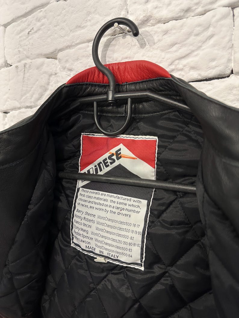 May include: Black leather jacket with a red collar, hanging on a black hanger. The jacket has a quilted black lining and a label with the brand name and text. The label also lists the names of World Champion riders.