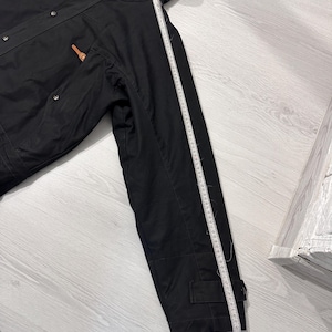 May include: A black hooded jacket with brown leather-like toggles and metal snap buttons. A white measuring tape is placed along the sleeve, showing a length of approximately 26 inches from the shoulder seam to the cuff edge of the garment.