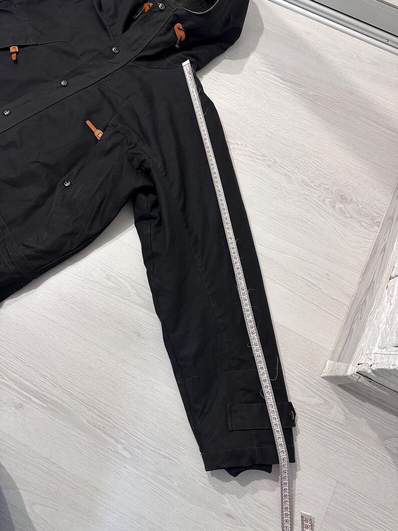 May include: A black hooded jacket with brown leather-like toggles and metal snap buttons. A white measuring tape is placed along the sleeve, showing a length of approximately 26 inches from the shoulder seam to the cuff edge of the garment.