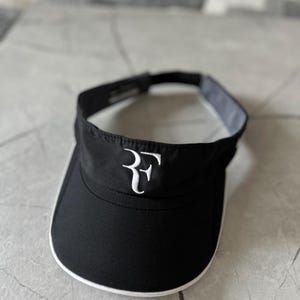 Può includere: Visiera nera con finiture bianche e le iniziali bianche ricamate "RF" sul davanti. La visiera ha una visiera curva e una fascia interna grigio scuro. Progettata per proteggere gli occhi dal sole.