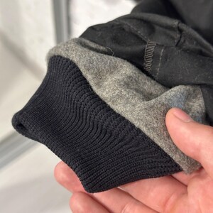 May include: A black jacket sleeve featuring a ribbed black knit cuff and a grey felted wool lining. The outer shell is constructed from a solid black fabric with visible stitching along the seam where the cuff meets the sleeve material.