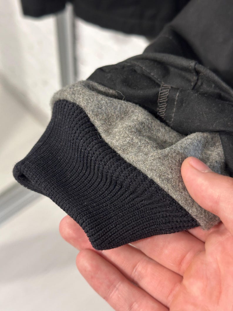 May include: A black jacket sleeve featuring a ribbed black knit cuff and a grey felted wool lining. The outer shell is constructed from a solid black fabric with visible stitching along the seam where the cuff meets the sleeve material.