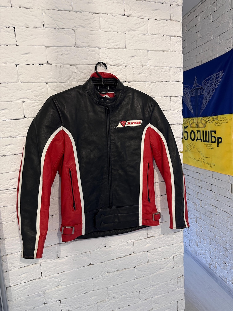 May include: Black leather motorcycle jacket with red and white accents. The jacket features a zip-up front, a small logo on the chest, and a buckled waist. The sleeves have white stripes and red panels.