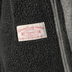May include: A dark charcoal gray fleece jacket with a textured, fuzzy surface. A white rectangular label is stitched onto the chest, featuring red text that reads: A GENUINE FILSON GARMENT, C.C. FILSON CO. SEATTLE, MIGHT AS WELL HAVE THE BEST.