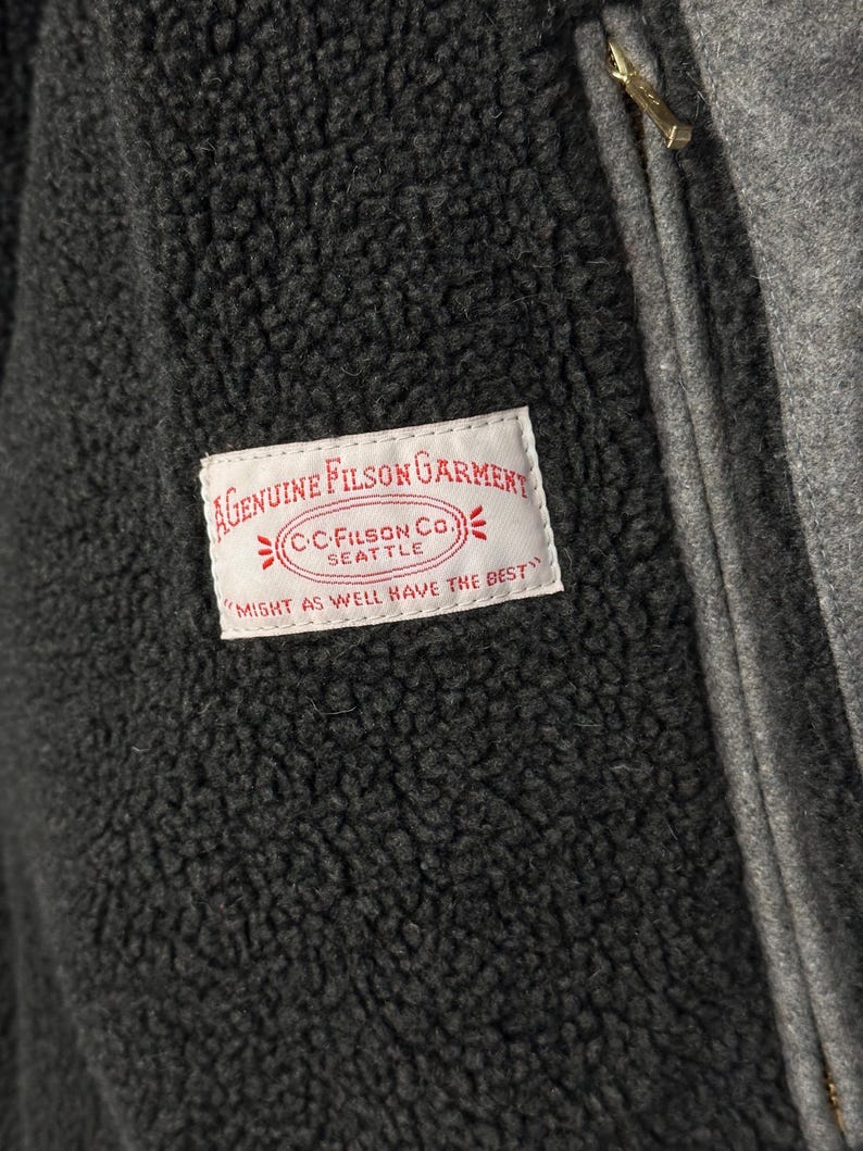 May include: A dark charcoal gray fleece jacket with a textured, fuzzy surface. A white rectangular label is stitched onto the chest, featuring red text that reads: A GENUINE FILSON GARMENT, C.C. FILSON CO. SEATTLE, MIGHT AS WELL HAVE THE BEST.