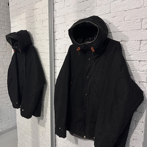 May include: A black hooded jacket featuring a front zipper closure, snap buttons, and brown leather toggles on the hood drawstrings. The jacket has long sleeves with adjustable cuffs and a structured hood, providing a solid black outer layer.