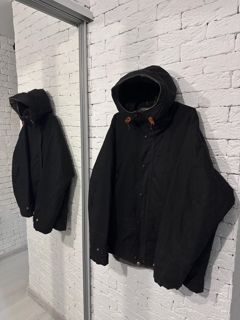 May include: A black hooded jacket featuring a front zipper closure, snap buttons, and brown leather toggles on the hood drawstrings. The jacket has long sleeves with adjustable cuffs and a structured hood, providing a solid black outer layer.