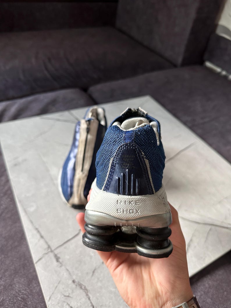 May include: A pair of Nike Shox athletic shoes. The shoes are primarily navy blue and white, with a mesh upper and a distinctive Shox sole. The Nike Shox logo is visible on the heel.