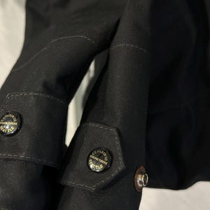 May include: A black jacket featuring long sleeves with adjustable cuffs. Each cuff is secured by a black button embossed with the text C.C. FILSON CO. MANUFACTURERS SEATTLE. The fabric has a visible weave texture and includes a metal snap fastener.
