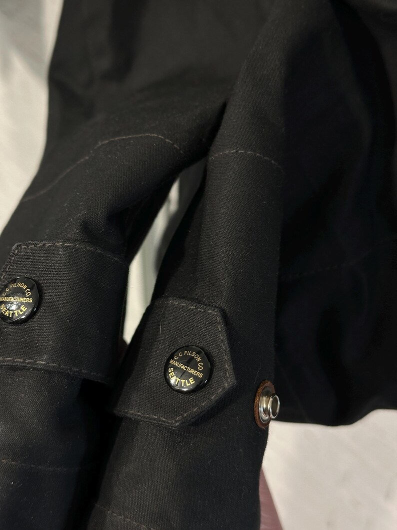 May include: A black jacket featuring long sleeves with adjustable cuffs. Each cuff is secured by a black button embossed with the text C.C. FILSON CO. MANUFACTURERS SEATTLE. The fabric has a visible weave texture and includes a metal snap fastener.