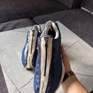 May include: A pair of blue and beige sneakers with a zipper closure. The sneakers have a mesh and leather design, with a white sole. The shoes are on a marble surface.