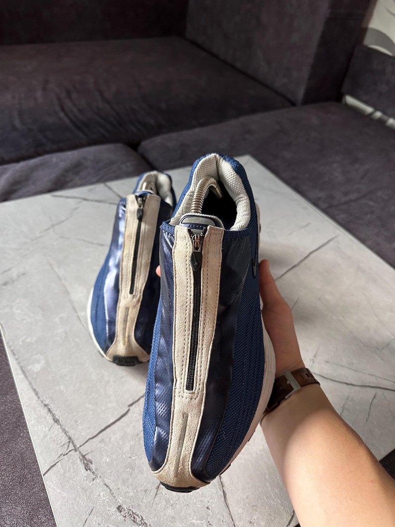 May include: A pair of blue and beige sneakers with a zipper closure. The sneakers have a mesh and leather design, with a white sole. The shoes are on a marble surface.