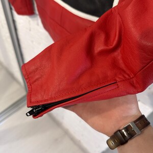 May include: Close-up of a red leather jacket with black and white accents. The jacket features a zippered sleeve cuff. The leather appears to be smooth and the stitching is precise. The jacket is likely part of a motorcycle or racing apparel collection.
