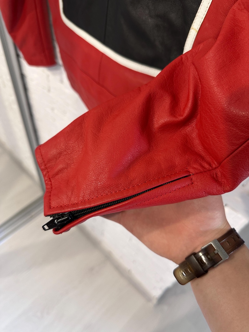 May include: Close-up of a red leather jacket with black and white accents. The jacket features a zippered sleeve cuff. The leather appears to be smooth and the stitching is precise. The jacket is likely part of a motorcycle or racing apparel collection.