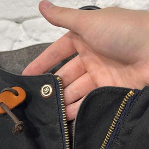 May include: A dark charcoal gray jacket featuring a brass-colored metal zipper closure, a matching brass snap button, and a brown leather pull tab attached to a dark brown cord. The garment has a dark blue fabric trim along the zipper edge.