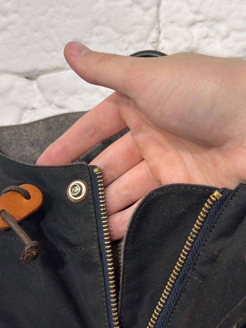 May include: A dark charcoal gray jacket featuring a brass-colored metal zipper closure, a matching brass snap button, and a brown leather pull tab attached to a dark brown cord. The garment has a dark blue fabric trim along the zipper edge.