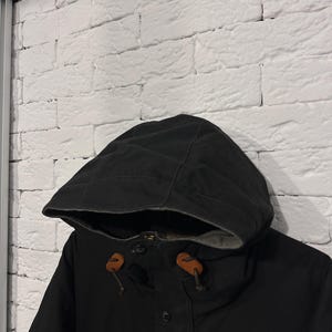 May include: A black hooded jacket featuring a dark grey interior lining, a front button closure, and two brown leather toggles attached to the hood drawstrings. The jacket is constructed from a solid black fabric with visible stitching details.