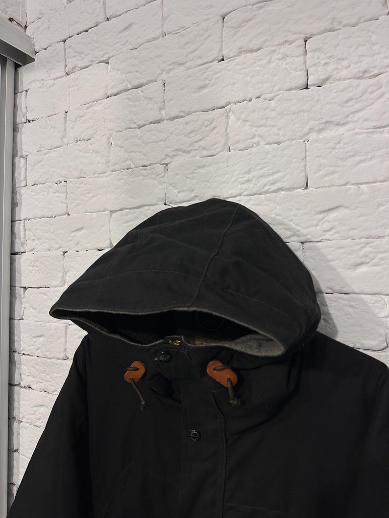 May include: A black hooded jacket featuring a dark grey interior lining, a front button closure, and two brown leather toggles attached to the hood drawstrings. The jacket is constructed from a solid black fabric with visible stitching details.
