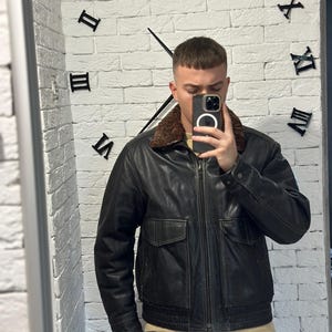 May include: Black leather jacket with a brown fur collar, two front pockets, and a zipper closure. The jacket is worn over a light-colored sweater. The background features a white brick wall with a large clock.