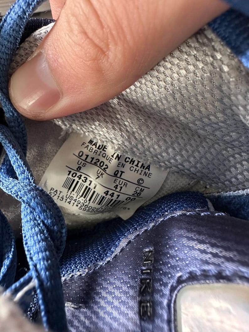 May include: Close-up of the inside of a blue and white Nike sneaker. The label reads "MADE IN CHINA" with size information: US 8, UK 7, EUR 41, and CM 26. The shoe's interior is a textured gray fabric.