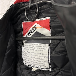 May include: Black leather motorcycle jacket with a red collar, hanging on a metal hanger. The interior features a quilted black lining and a tag with the brand logo and text. The jacket is size 44 and made in Italy.