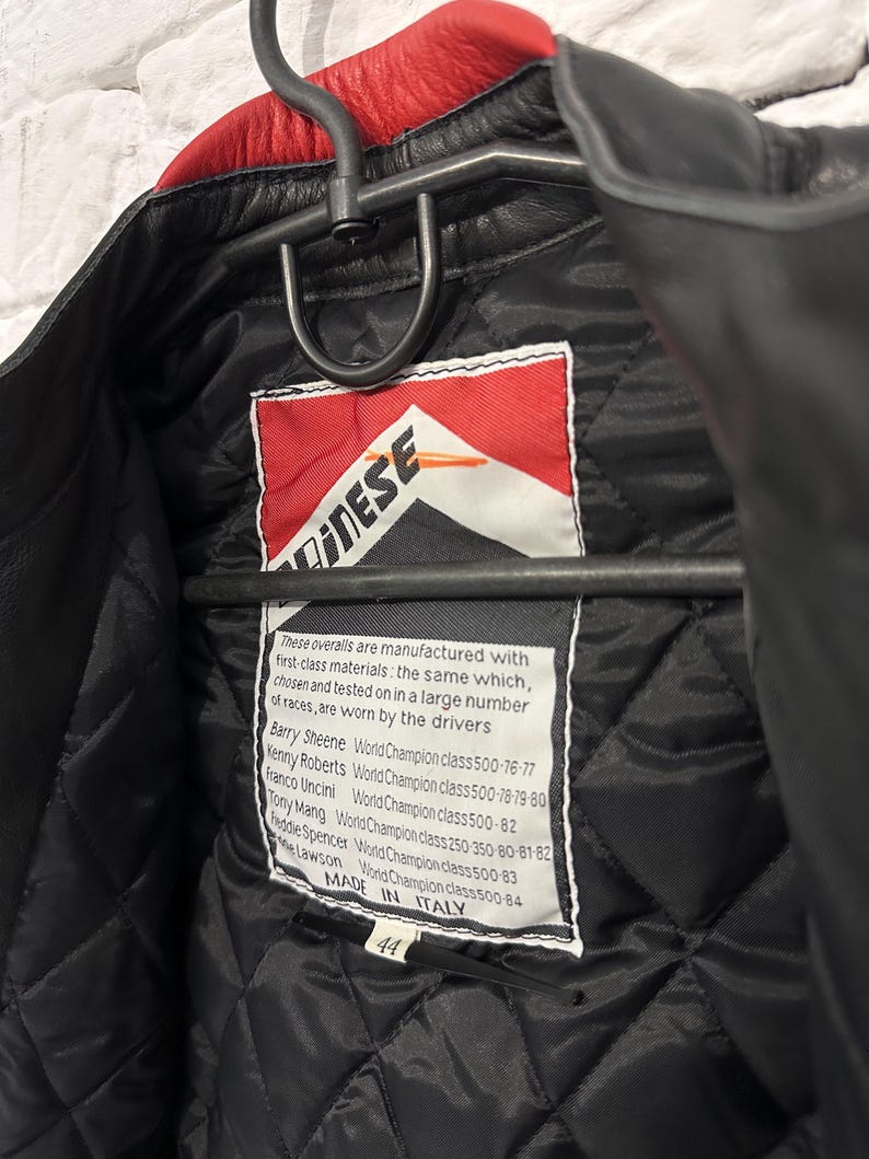 May include: Black leather motorcycle jacket with a red collar, hanging on a metal hanger. The interior features a quilted black lining and a tag with the brand logo and text. The jacket is size 44 and made in Italy.