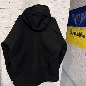 May include: A solid black hooded jacket featuring long sleeves, a structured hood, and a horizontal seam detail across the back. The garment is displayed from the rear view, showcasing a plain, dark fabric construction with clean lines and stitching.
