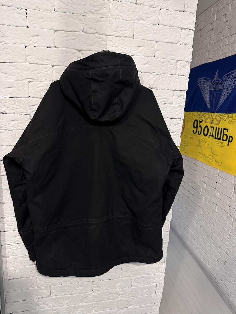 May include: A solid black hooded jacket featuring long sleeves, a structured hood, and a horizontal seam detail across the back. The garment is displayed from the rear view, showcasing a plain, dark fabric construction with clean lines and stitching.