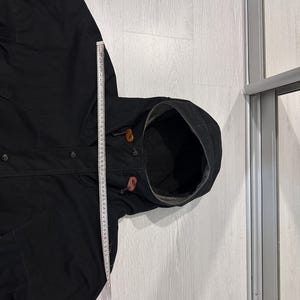 May include: A black hooded jacket with a dark interior lining, featuring brown leather toggles on the hood drawstrings and a front button closure. A white measuring tape is placed vertically along the side, showing a length of 60 centimeters.