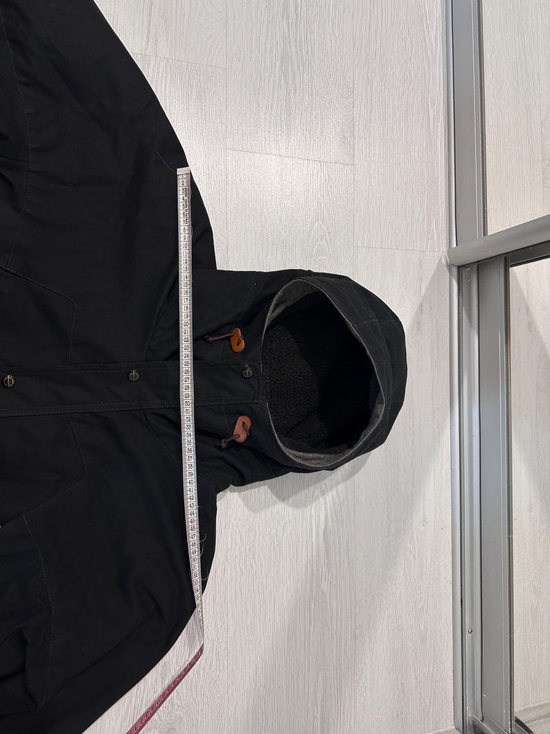 May include: A black hooded jacket with a dark interior lining, featuring brown leather toggles on the hood drawstrings and a front button closure. A white measuring tape is placed vertically along the side, showing a length of 60 centimeters.