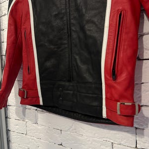 May include: Black and red leather jacket with white accents. The jacket features a central black panel, red side panels, and white stripes. It has a zippered front, pockets, and adjustable waist straps.