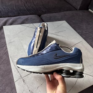 May include: A pair of blue and white athletic shoes with a zipper closure. The shoes have a mesh upper, a black Nike swoosh, and a unique sole design with black and gray cushioning. The shoes are displayed on a gray surface.