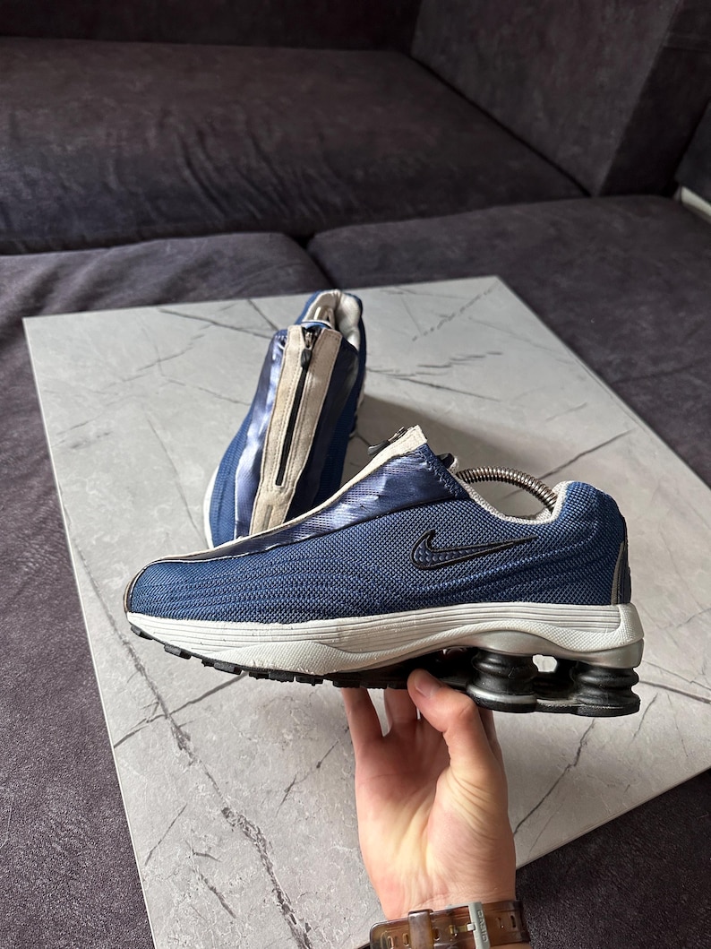May include: A pair of blue and white athletic shoes with a zipper closure. The shoes have a mesh upper, a black Nike swoosh, and a unique sole design with black and gray cushioning. The shoes are displayed on a gray surface.