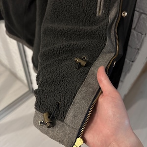 May include: A black jacket with a dark gray sherpa fleece lining. The jacket features a brass-colored metal zipper and two brass-colored toggle fasteners with black cords attached to a gray fabric trim along the front opening edge.