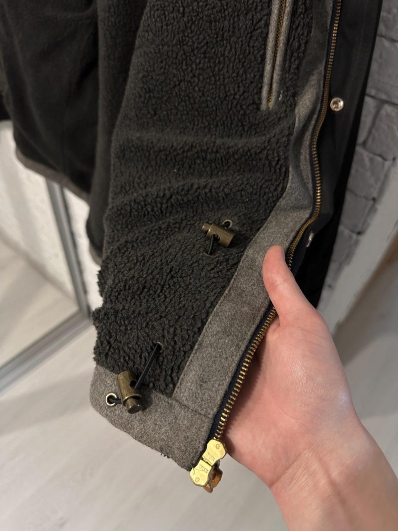 May include: A black jacket with a dark gray sherpa fleece lining. The jacket features a brass-colored metal zipper and two brass-colored toggle fasteners with black cords attached to a gray fabric trim along the front opening edge.