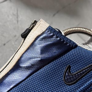 May include: Close-up of a blue and white athletic shoe. The shoe features a blue mesh fabric, a metallic blue accent, and a black Nike swoosh logo. A zipper and a silver metal detail are also visible.