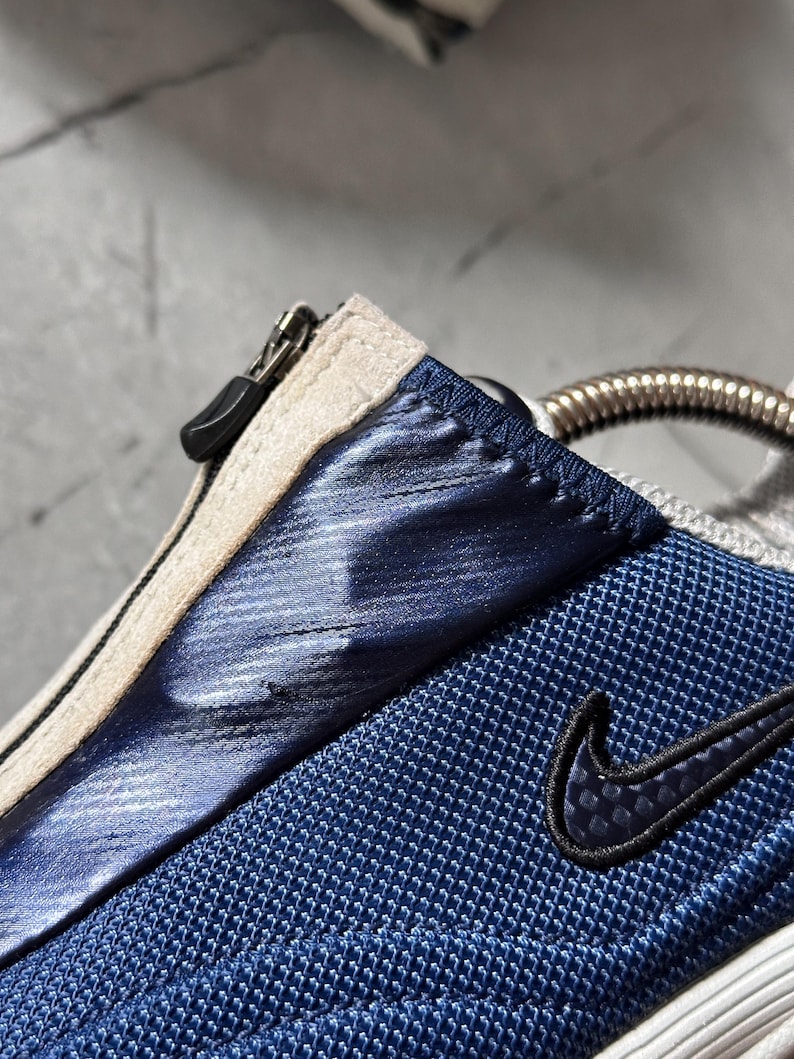 May include: Close-up of a blue and white athletic shoe. The shoe features a blue mesh fabric, a metallic blue accent, and a black Nike swoosh logo. A zipper and a silver metal detail are also visible.
